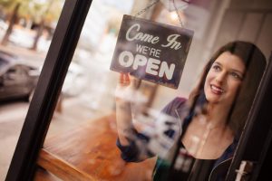 Hipster woman smiling and looking out the window, turns opening sign on front door of coffee shop with reflection of street in glass