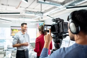 Happy businessman during corporate interview with female journalist. Manager answering question in office. Young woman at work as reporter with business man and cameraman shooting video for broadcast