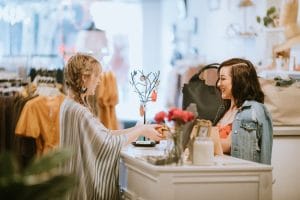 One cashier serving a customer in a boutique over the counter. In the background there is a clothes on hanging racks for sale. 