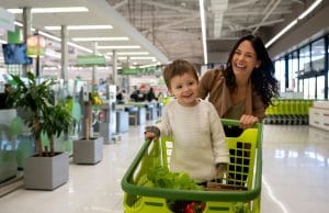 Mother pushing cart with her toddler inside with purchased groceries leaving the supermarket and facing camera smiling very excited