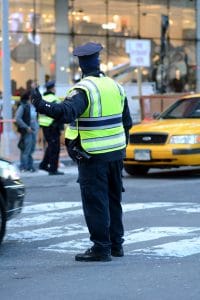 Police office standing in the road directing traffic between a yellow taxi on his right and a black car on his left 