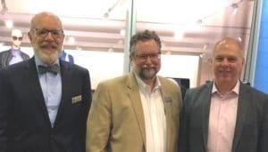 Three men standing up next to each other, wearing business suits and smiling towards the camera.